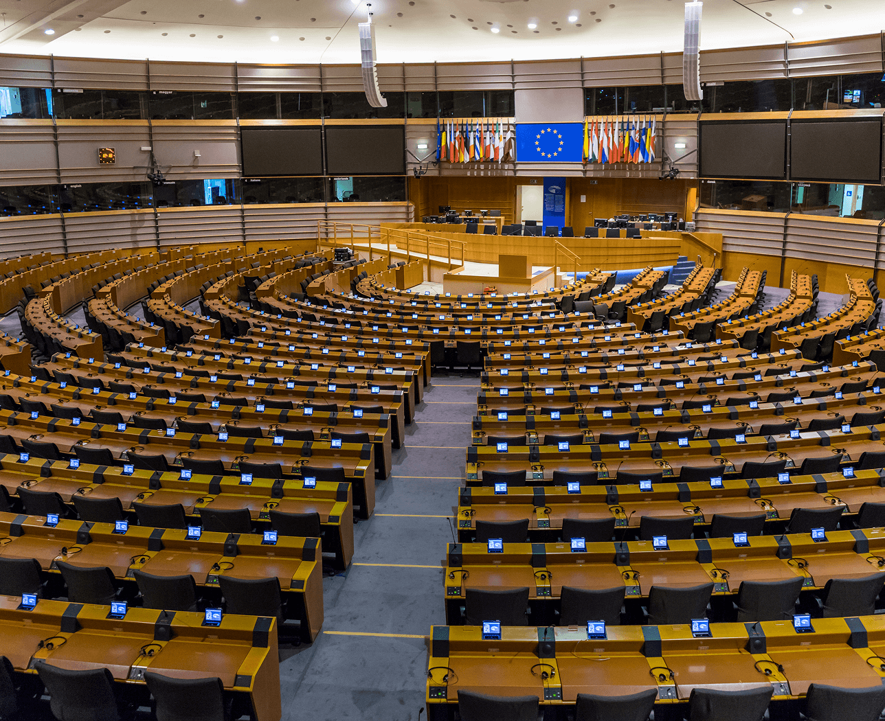 Empty European Parliament chamber with curved rows of desks and chairs facing a speaker's podium and flags.