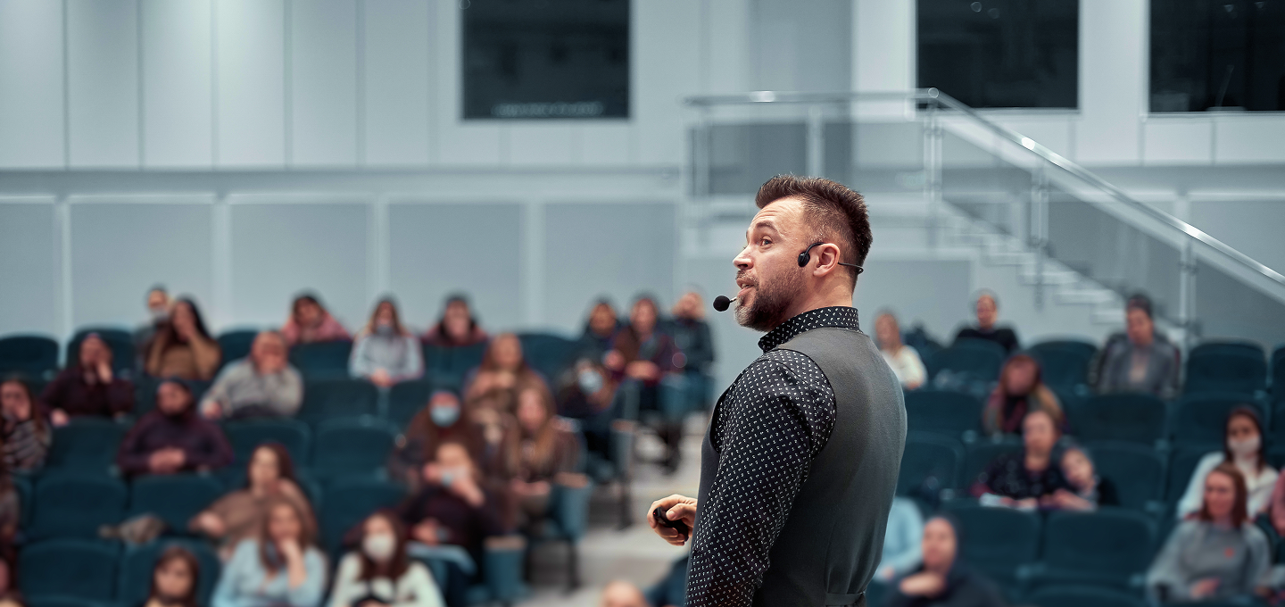 Speaker wearing a microphone headset presenting to a seated audience in a lecture hall.